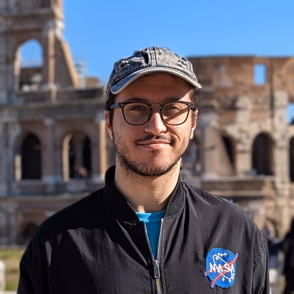 Adam (Last Level Studios Director) stood in front of Rome's Colosseum, wearing a grey baseball cap, glasses and a NASA jacket.